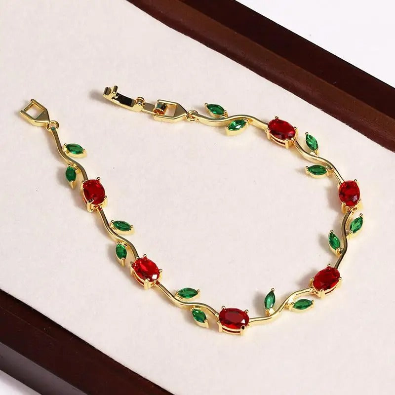 A gold vine-style bracelet with oval red gemstones and small green leaf accents, displayed on a cream surface with dark brown borders. sasha jewels 