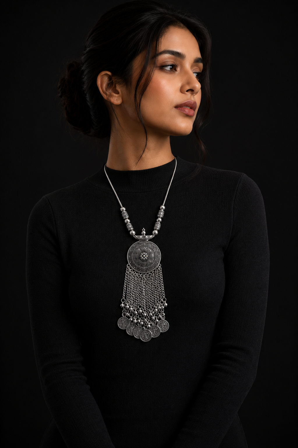 A woman wearing a large silver-toned pendant necklace with dangling chain and coin-like charms, photographed against a black background. sasha jewels / jewellery / necklaces 